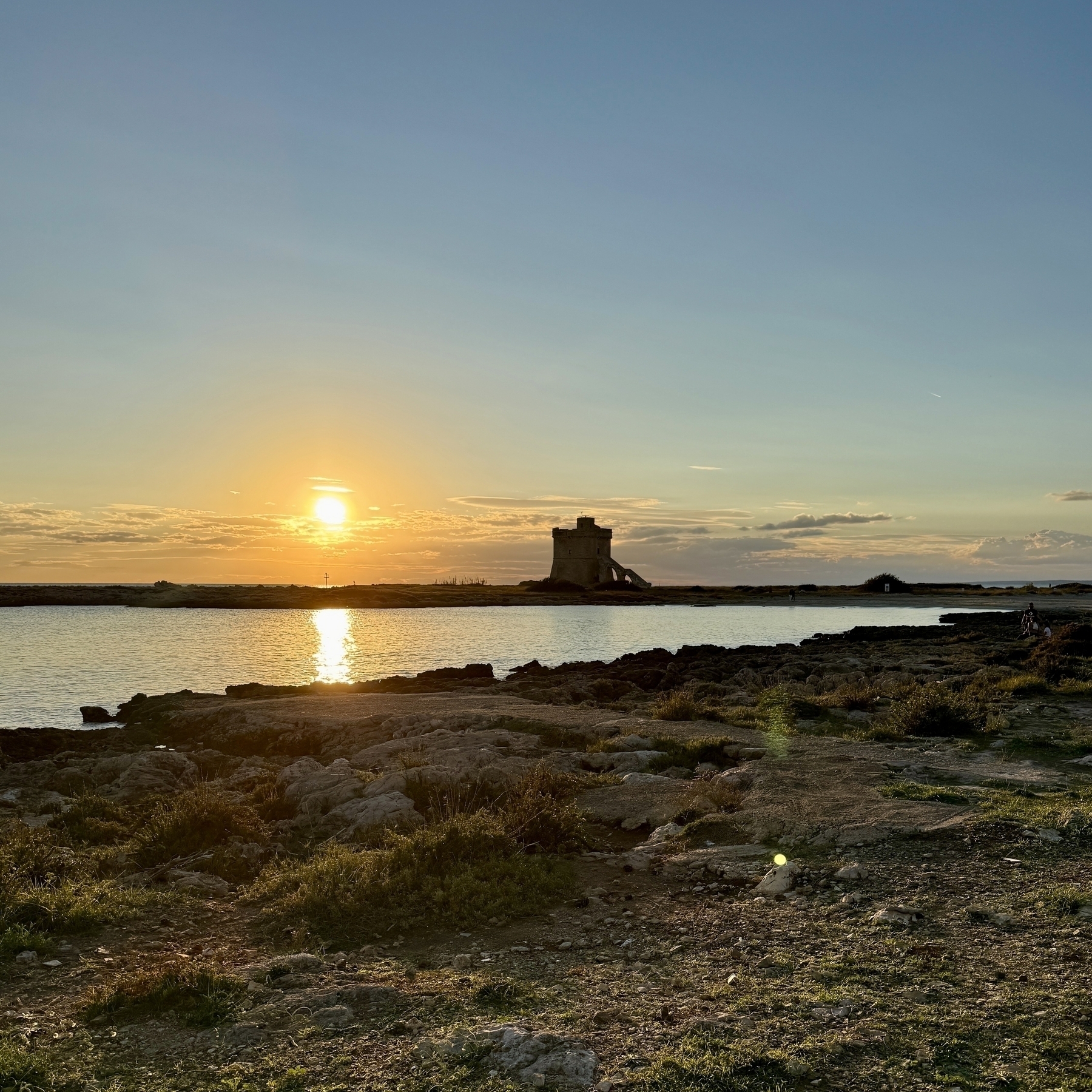 A sunset at a beach in Italy, the sky is blue above the ocean and there are rocks with green grass.