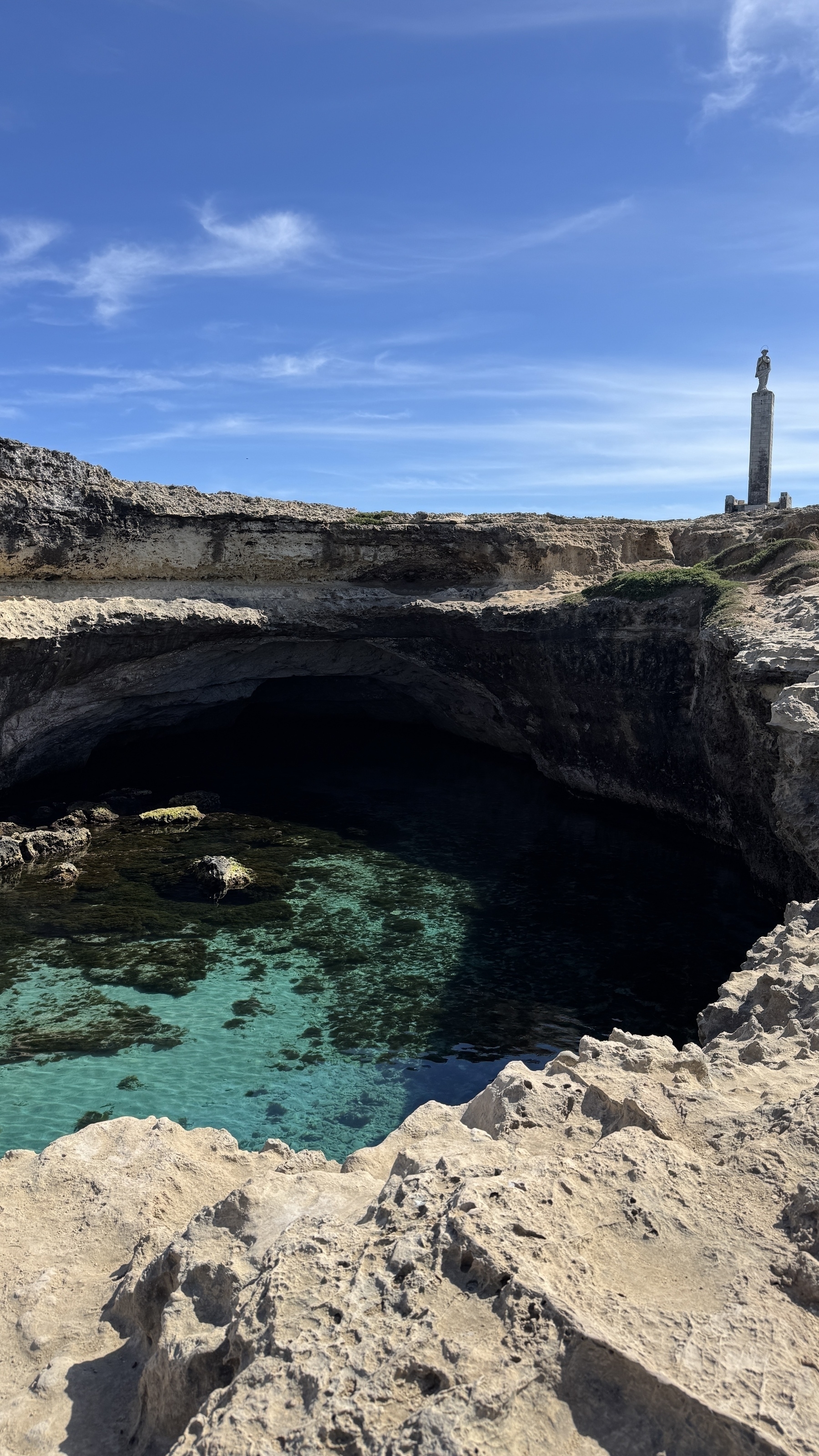 Grotta della Poesia in Salento. It is a circular pool of seawater surrounded by cliffs. The sky is clear and blue and the water in the pool is light blue.